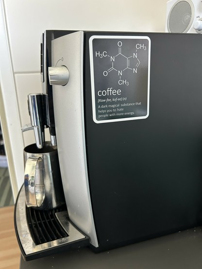 An espresso machine with a portafilter in place, and a humorous sticker on the front showing the chemical structure of caffeine and a playful definition of coffee.