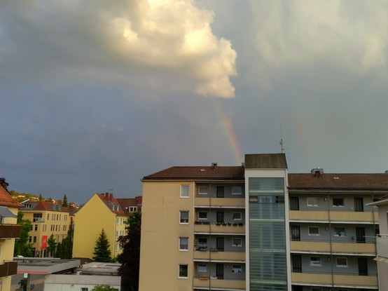 Ein kleiner Regenbogen, der sich in Wolken verliert, ist hinter einem Häuserblock zu sehen.